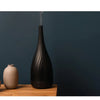 Made by Zen Thalia Dusk Diffuuseri; korkea, musta ultraäänidiffuusori mattapinnalla päästää ohuen sumuvirran; diffuusori modernin lieriömäinen ilman näkyvää etikettiä tai tekstiä; sijoitettuna puupinnalle pienen, pyöreähkön beigen keraamisen maljakon viereen; taustalla yhtenäinen tummansininen seinä; ei käsiä tai ihmisiä kuvassa.
