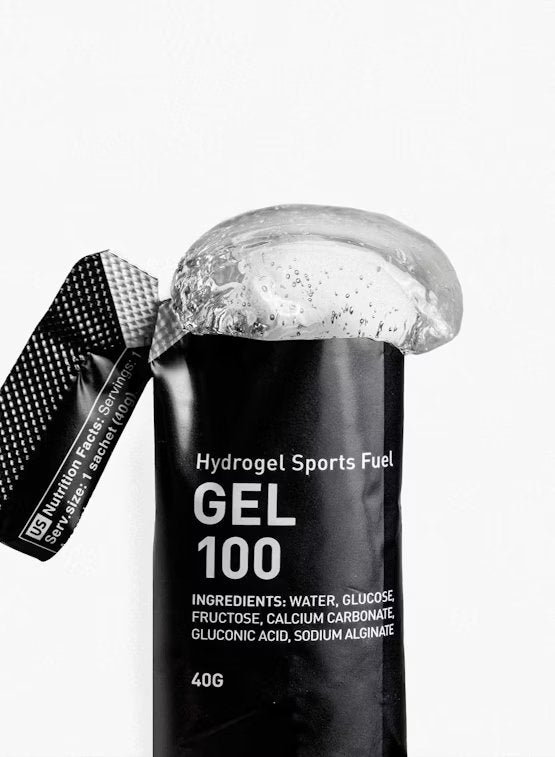 Maurten Gel 100 (12-pack), musta urheilugeelipussi merkitty "Hydrogel Sports Fuel GEL 100", geeliä valumassa ulos; pakkaus mustaa mattapintaista, joustava suorakulmainen muoto; ainesosat ja ravintosisältötiedot painettu selkeästi pienellä fontilla takaosaan; pääetiketti käyttää groteskityppografiaa; tausta valkoinen, lähikuva tuotteesta, ei ihmisiä eikä käsiä näkyvissä.