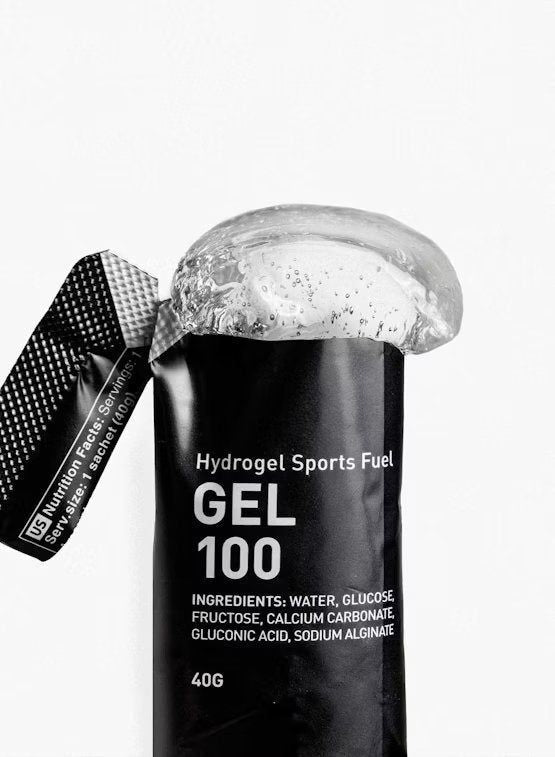 Maurten Gel 100 (12-pack), musta urheilugeelipussi merkitty "Hydrogel Sports Fuel GEL 100", geeliä valumassa ulos; pakkaus mustaa mattapintaista, joustava suorakulmainen muoto; ainesosat ja ravintosisältötiedot painettu selkeästi pienellä fontilla takaosaan; pääetiketti käyttää groteskityppografiaa; tausta valkoinen, lähikuva tuotteesta, ei ihmisiä eikä käsiä näkyvissä.