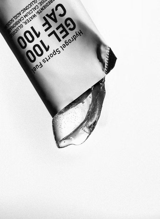 Maurten Gel 100 CAF 100 (12-pack); lähikuva osittain avatusta urheilugeelipussista, kirkas geeli valumassa ulos valkoista taustaa vasten; pussi suorakulmainen lovetuin reunoin, kiiltävää joustavaa materiaalia ja painetuin musta-valkoisin tekstein lihavoidulla groteskifontilla; etiketissä lukee GEL 100 CAF 100 sekä muut ravintosisältötiedot; ei ihmisiä eikä käsiä näkyvissä.