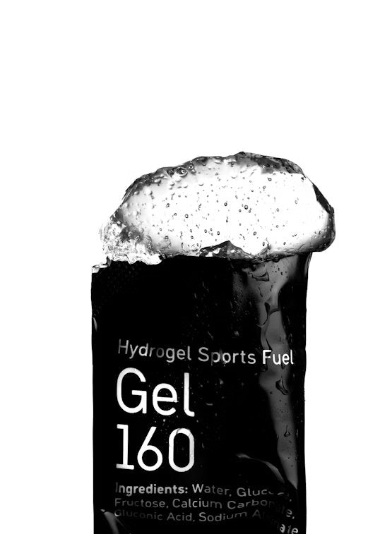 Maurten Gel 160 (12-pack): musta suorakulmainen pakkaus mattapinnalla tekstillä "Hydrogel Sports Fuel Gel 160" lihavoidulla groteskikirjasimella; vaahtoavaa geeliä valuu näkyvästi pakkauksen yläreunan avauksesta; osa ainesosien nimistä on luettavissa pakkauksen alaosassa; tuote yhtenäistä valkoista taustaa vasten lähikuvassa; ei ihmisiä tai käsiä näkyvissä.