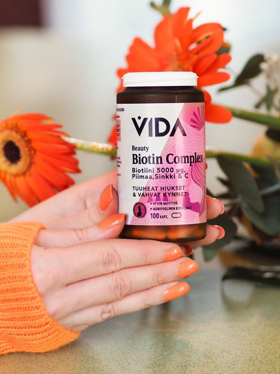 Vida Beauty Biotin Complex; oransseilla kynsillä maalattu käsi pitelee lieriön muotoista, läpinäkymätöntä valkoista pulloa VIDA Beauty Biotin Complex -tuotteelle mattapinnalla ja valkoisella korkilla; etiketti painettu tummalla sans-serif-tekstillä listaten biotiinin, sinkin ja tärkeät ravintoaineet; tausta täynnä kirkkaanoransseja kukkia; lähikuva.
