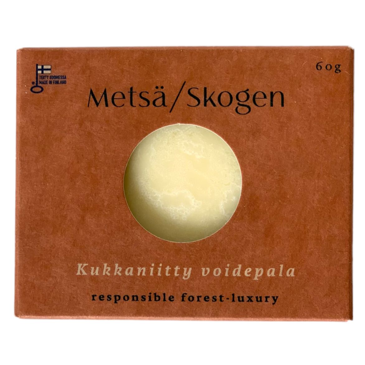 A brown matte-finish rectangular box labeled Metsä/Skogen features a circular transparent window showing a round, solid cream-colored item inside. The box has legible text: Kukkaniitty voidepala, responsible forest-luxury, and 60g in simple sans-serif font. No people or hands are visible. Neutral background with minimal context shown.