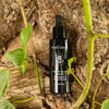 Moi Forest Glow Serum; musta tippapullo lepää puunoksalla vihreiden lehtien, sammaleen ja adaptogeenisten kasvien ympäröimänä, sulautuen luonnolliseen ulkoympäristöön; pullo sylinterimäinen mattapinnalla, etiketissä valkoista groteskitypografiaa; ei ihmisiä eikä käsiä näkyvissä; lähikuva tuotteesta ulkotiloissa.