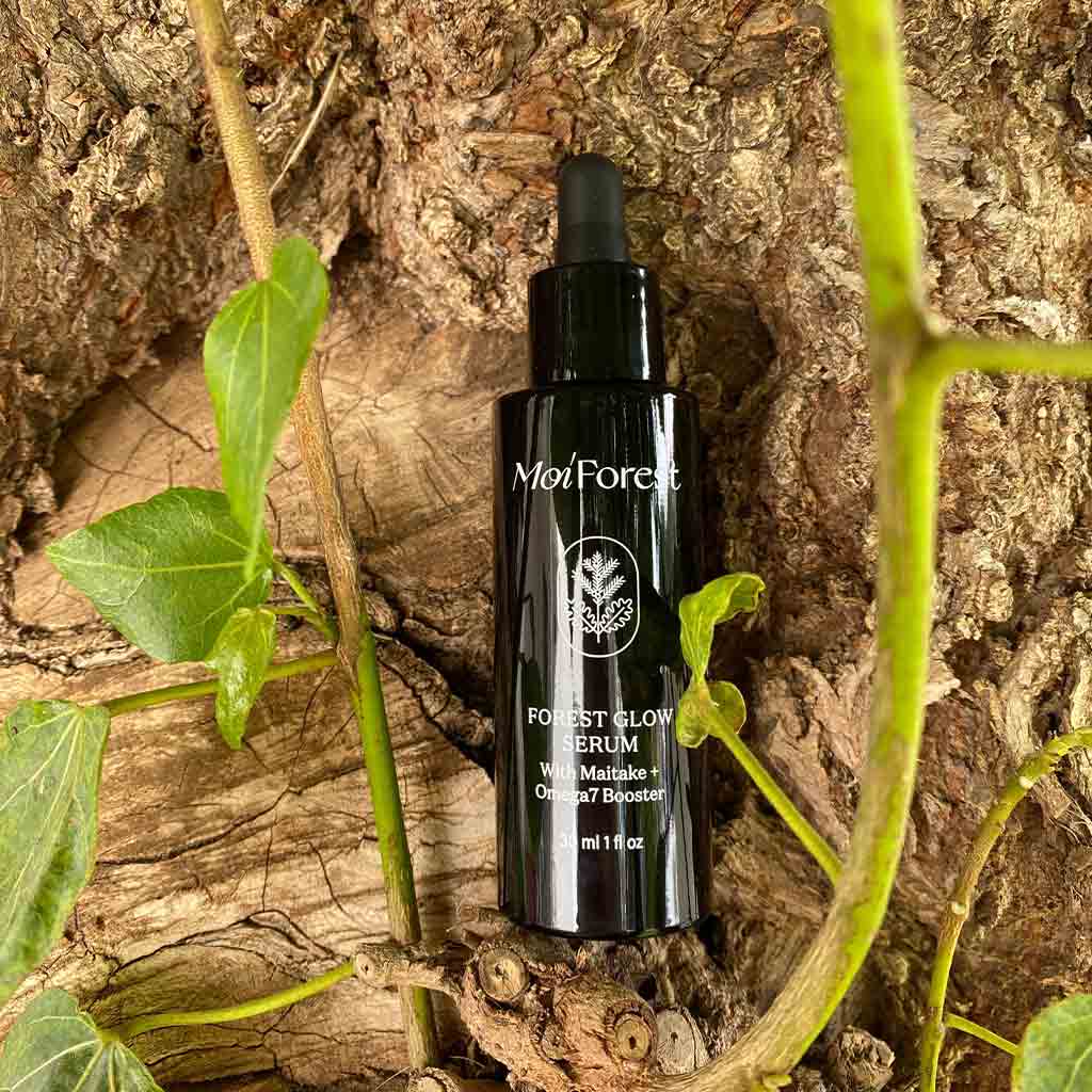 Moi Forest Glow Serum; musta tippapullo lepää puunoksalla vihreiden lehtien, sammaleen ja adaptogeenisten kasvien ympäröimänä, sulautuen luonnolliseen ulkoympäristöön; pullo sylinterimäinen mattapinnalla, etiketissä valkoista groteskitypografiaa; ei ihmisiä eikä käsiä näkyvissä; lähikuva tuotteesta ulkotiloissa.