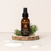 Moi Forest Rain Face Mist; ruskea mattapintainen suihkepullo kirkkaalla korkilla ja valkoisella etiketillä tekstillä "MoiForest Forrest Rain Face Mist" yksinkertaisella sans-serif-fontilla, pystyssä pyöreällä puuviipaleella; tummanvihreitä havunoksia aseteltu pullon viereen; vesipisaroita näkyvissä sileällä vaaleanbeigellä taustapinnalla; ei ihmisiä tai käsiä kuvassa.