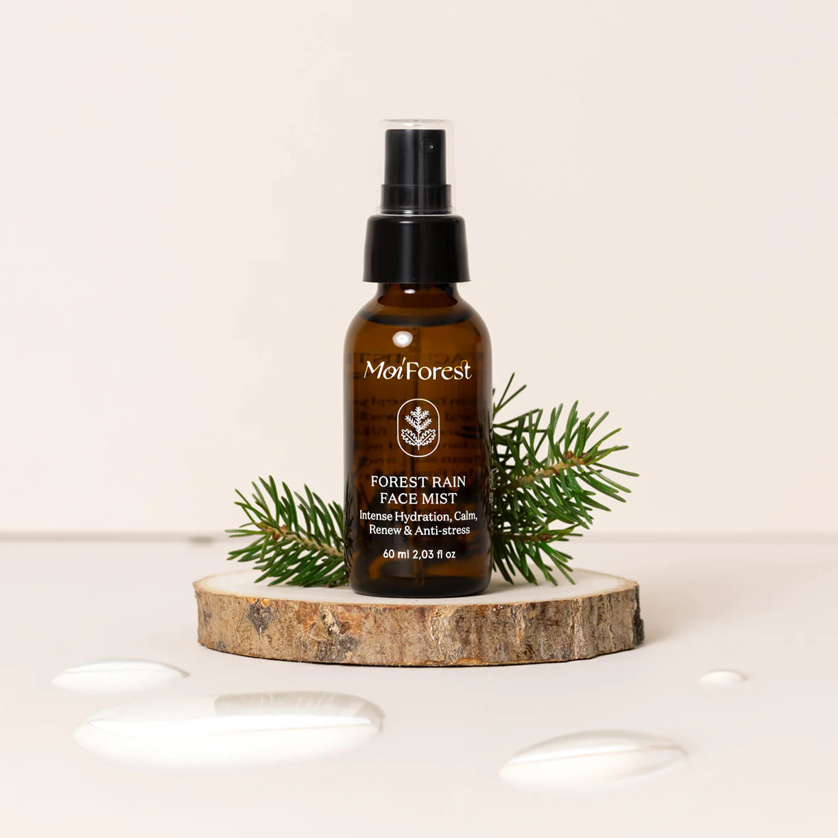 Moi Forest Rain Face Mist; ruskea mattapintainen suihkepullo kirkkaalla korkilla ja valkoisella etiketillä tekstillä "MoiForest Forrest Rain Face Mist" yksinkertaisella sans-serif-fontilla, pystyssä pyöreällä puuviipaleella; tummanvihreitä havunoksia aseteltu pullon viereen; vesipisaroita näkyvissä sileällä vaaleanbeigellä taustapinnalla; ei ihmisiä tai käsiä kuvassa.