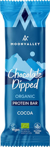 Moonvalley Luomu Proteiinipatukka Kaakao; sininen Moonvalley Chocolate Dipped Organic Protein Bar -kääre, kaakao-maku; suorakulmainen matta muovipakkaus valkoisella ja kultaisella tekstillä näyttää tuotenimen ja maun; etupuolella vegaani-, EU-luomu- ja gluteenittomuuskuvakkeet sekä tyylitelty valkoinen vuorigrafiikka taustalla; ei ihmisiä tai muita esineitä, tasainen tausta, lähikuva.