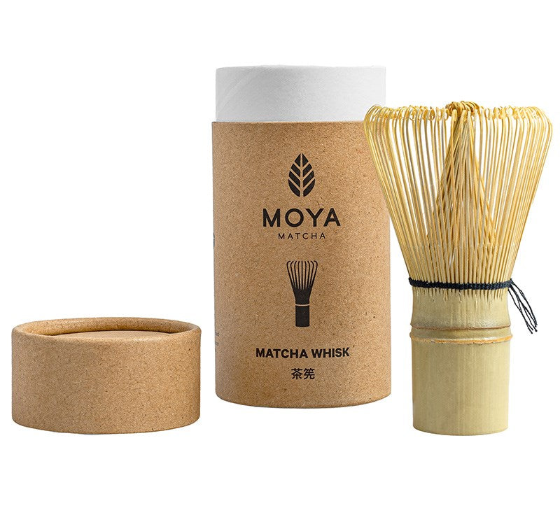 Moya Matcha Matchavispilä; bambuinen matchavispilä lieriömäisen ruskean irtokantisen purkin vieressä, purkissa teksti MOYA Matcha ja vispilän kuvitus; purkin pinta mattapintainen, teksti isoin groteskikirjaimin, kuvitus viivapiirroksena; molemmat vaalealla alustalla hyvin valaistussa tuotekuvassa; ei ihmisiä tai käsiä.