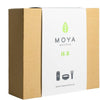 Brown corrugated box with a white label that says MOYA MATCHA. White matte rectangular label on front has sans-serif black text, a simple green leaf logo, and black outlined icons of a matcha tin, bowl, and whisk. No people or hands present. Plain background, product centered in frame. Tabletop surface visible under the box.