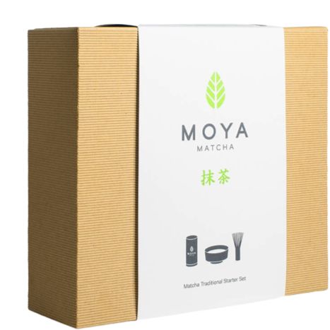 Brown corrugated box with a white label that says MOYA MATCHA. White matte rectangular label on front has sans-serif black text, a simple green leaf logo, and black outlined icons of a matcha tin, bowl, and whisk. No people or hands present. Plain background, product centered in frame. Tabletop surface visible under the box.