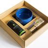 A boxed matcha tea set featuring a blue ceramic bowl with a matte finish, a metal tin labeled “Moya Matcha,” and a natural bamboo whisk, all arranged inside a rectangular brown cardboard box with divider compartments. White sans-serif typography on the tin is visible. Background is plain white. No people or hands are shown.