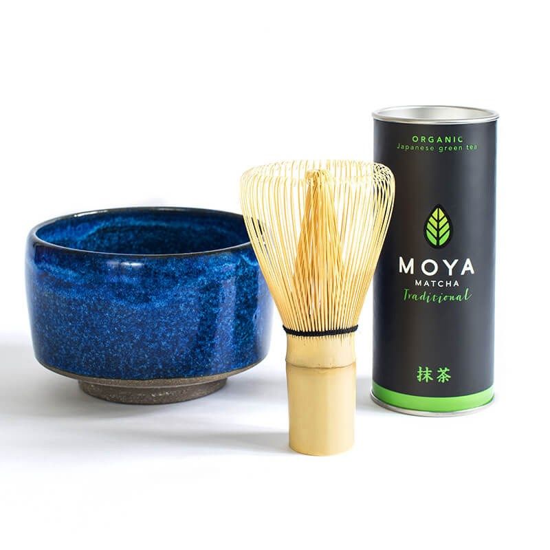 A blue ceramic matcha bowl with a matte finish sits next to a cylindrical bamboo matcha whisk and a glossy metal canister labeled “Moya organic matcha tea” in bold sans-serif type. All objects are arranged on a plain white background. No people or hands are visible. Shot is directly overhead; all products fully visible.