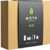 A brown rectangular box with a matte finish, featuring a black label on the front. The label has the MOYA Matcha logo in bold white font, a green leaf graphic, Japanese text, and small icons depicting premium matcha tea accessories. Typography includes clean sans-serif and stylized Japanese characters. Background is plain white; no people or hands shown. Box is the only item present.