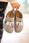 A child holds gray slippers featuring Moomin character graphics on the tops. The slippers appear soft with a matte fabric finish and have a rounded toe shape. The child wears pajama pants with a pattern of multicolored stars and crescent moons. No product packaging or text labels are visible in the image. The background is out of focus. A child holds gray slippers decorated with adorable Moomin characters, while wearing colorful pajama pants patterned with stars and moons.