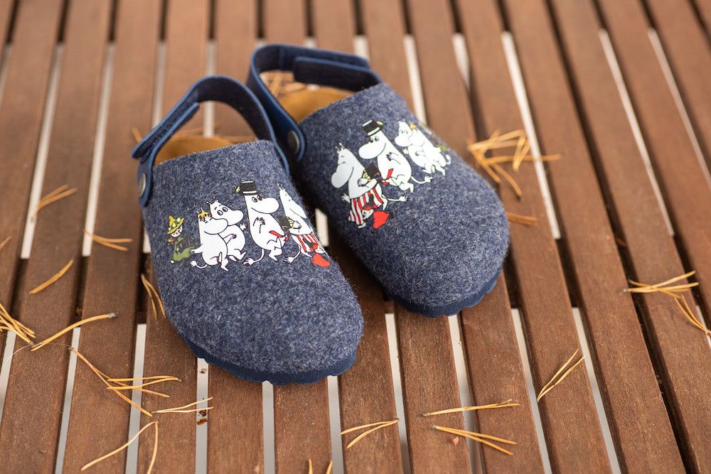 A pair of blue felt slippers featuring Moomin characters on top, resting on a wooden slatted surface with scattered pine needles. Slippers are rounded at the toe and do not show visible branding or packaging. The finish of the felt is matte. The Moomin characters appear as fabric patches with detailed outlines in muted colors. No hands, feet, or people present in the photo.