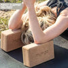 A person uses two rectangular cork yoga blocks with the word “myoga” printed on them to support their back and head while lying on a flat, solid-colored yoga mat outside. The surface of the blocks is matte. The photo is taken outdoors on grass. Simple sans-serif text in the image reads, Perfect to support restorative yoga poses.