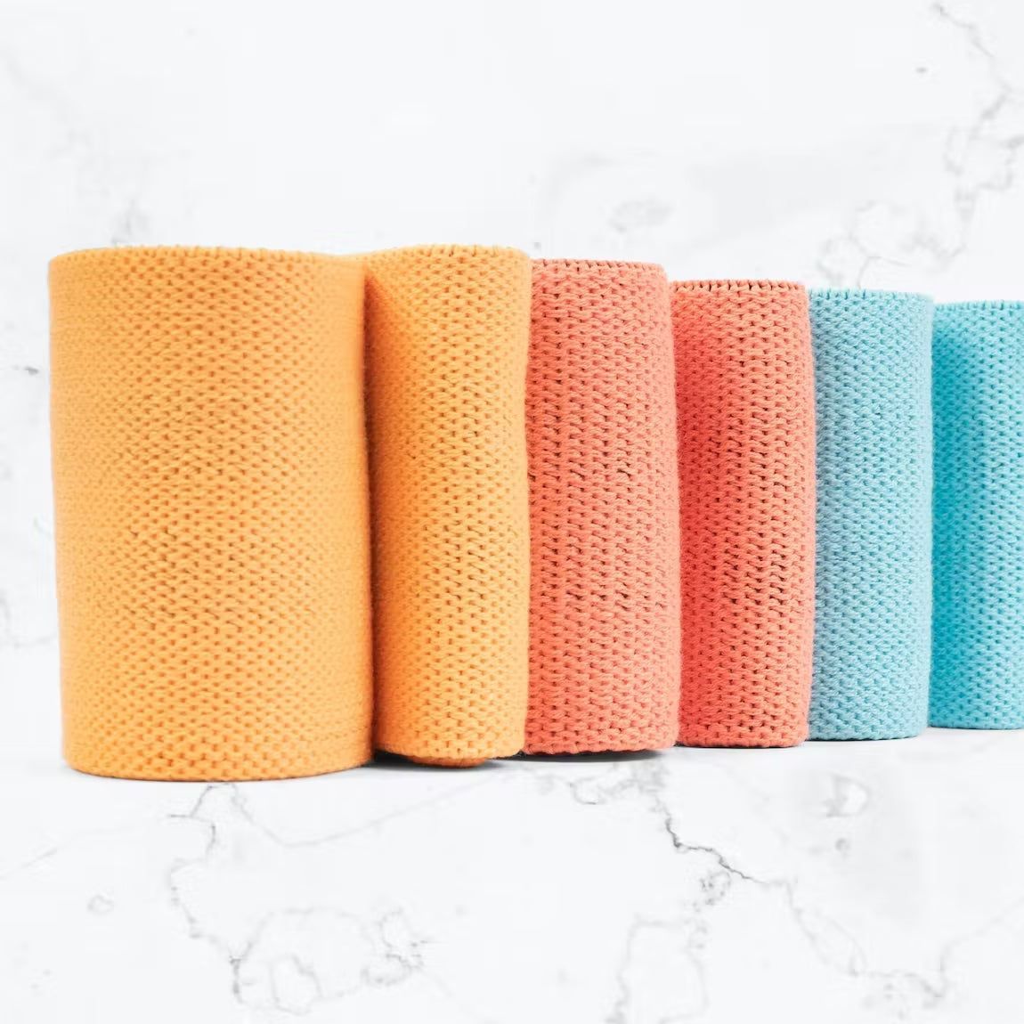 Myga vastusnauha 3-pack; viisi tekstuurista kangasrullaa oranssina, vaaleanpunaisena ja sinisenä pystyssä rivissä valkoisella marmoripinnalla, pyöristetyin yläreunoin; mattapintaiset kuvioidut pinnat; ei näkyvää pakkausta tai etikettejä; laaja kuva ylhäältä valaistuna.
