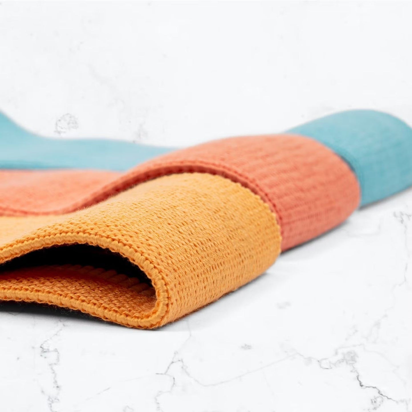 A close-up of a rolled, textured yoga mat featuring orange, coral, and teal stripes, placed on a white marble surface. The mat is cylindrical in shape and made from a patterned material with a matte finish. No packaging or typography is visible. There are no people or hands in the image. The background consists solely of smooth white marble.