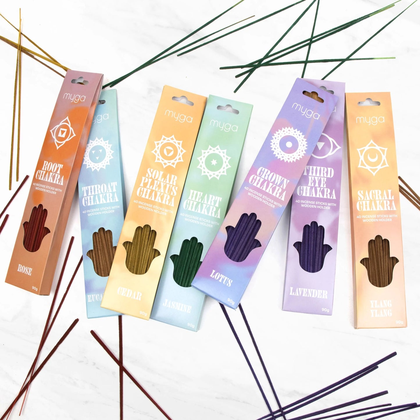 Myga Chakra Suitsuke; kuusi suorakulmaista chakra-aiheista suitsukerasiaa, kukin eri värinen ja merkitty vastaavan chakran nimellä ja tuoksulla selkeällä groteskikirjasimella, aseteltuna viuhkamaisesti mattavalkoiselle taustalle; useita irtonaisia tummia suitsuketikkuja on levitetty pakkausten ympärille; ei ihmisiä tai käsiä näkyvissä kuvassa.