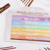 Myga Chakra Suitsukesetti; laatikko värikkäitä chakrasuitsuketikkuja, tuoksuina muun muassa lootus, laventeli, eukalyptus, jasmiini, setri, ylang ylang ja ruusu; suorakulmainen laatikko monivärisin osioin, kunkin tuoksun nimi valkoisella lihavoidulla groteskikirjasimella; laatikon vieressä mattapintainen puinen suitsuketeline ja useita irtonaisia sytyttämättömiä suitsuketikkuja neutraalilla taustalla; ei ihmisiä tai käsiä näkyvissä tuotekuvassa.
