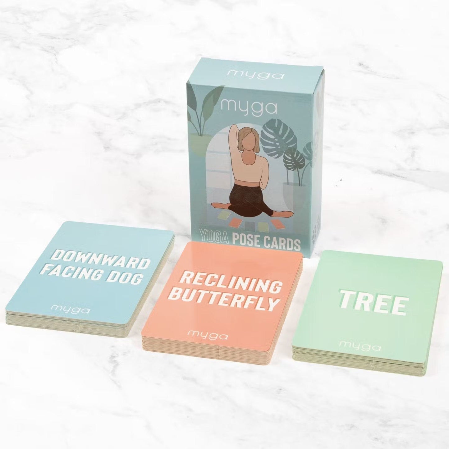 A box of Myga Yoga Pose Cards is displayed with three cards in front—Downward Facing Dog, Reclining Butterfly, and Tree—on a white marble surface. The box is rectangular, made of cardboard with a matte finish. Each card shows an illustrated yoga pose and pose name in clear sans-serif text. No people or hands are present. This is an overhead product shot against a smooth, patterned stone background.