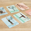 Yoga pose cards laid out on a wooden surface, each featuring an illustrated person in a different yoga position. Card backgrounds are white with color illustrations and black sans serif typography. Cards show the names Butterfly, Dangers Pose, Warrior Three, Headstand, and Side Stretch. Cards have square corners; no people or hands in frame. Overhead shot.