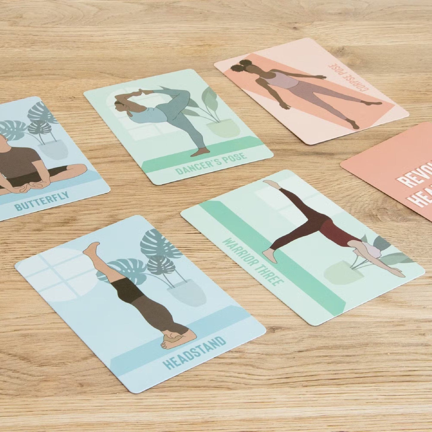 Yoga pose cards laid out on a wooden surface, each featuring an illustrated person in a different yoga position. Card backgrounds are white with color illustrations and black sans serif typography. Cards show the names Butterfly, Dangers Pose, Warrior Three, Headstand, and Side Stretch. Cards have square corners; no people or hands in frame. Overhead shot.