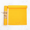 A bright yellow yoga mat is partially rolled out next to its rolled-up version, which features a brown paper label with printed yoga pose illustrations. Both mats are cylindrical in shape and have a matte finish. No typography is visible on the exterior of the mats or packaging. The products are positioned on a white marble surface with no people or hands present. The shot is directly overhead.