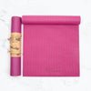 Two rectangular pink yoga mats made of textured material on a solid white surface; one mat is tightly rolled and held by a brown paper band, no visible graphics on the band. The second mat is partially unrolled showing an embossed, lowercase “myga” logo in simple sans-serif type. No people, props, or shadows are present. Studio product shot.