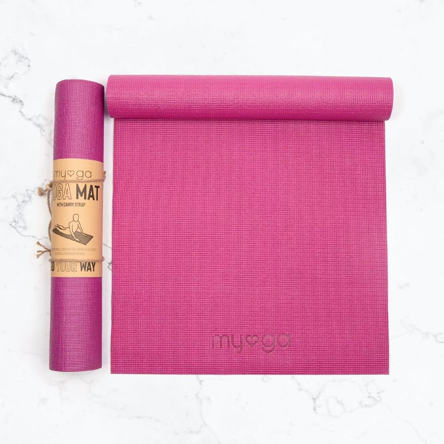 Two rectangular pink yoga mats made of textured material on a solid white surface; one mat is tightly rolled and held by a brown paper band, no visible graphics on the band. The second mat is partially unrolled showing an embossed, lowercase “myga” logo in simple sans-serif type. No people, props, or shadows are present. Studio product shot.