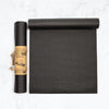 A black yoga mat lies unrolled beside a rolled-up version with a brown paper label reading MYOGA YOGA MAT, displayed on a white marble surface. Both mats are rectangular with rounded edges and have a matte finish. The brown label features large bold uppercase sans-serif text. There is no visible packaging other than the label, and no people or hands are present in the image. The shot is overhead.