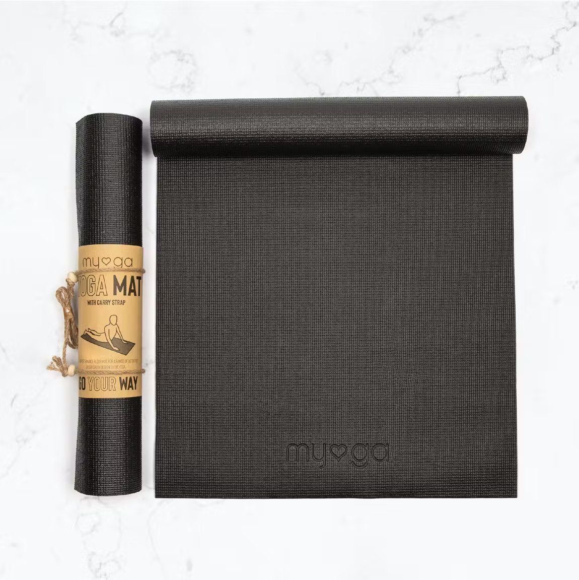 A black yoga mat lies unrolled beside a rolled-up version with a brown paper label reading MYOGA YOGA MAT, displayed on a white marble surface. Both mats are rectangular with rounded edges and have a matte finish. The brown label features large bold uppercase sans-serif text. There is no visible packaging other than the label, and no people or hands are present in the image. The shot is overhead.