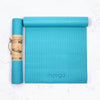 A rolled and an unrolled turquoise yoga mat on a white surface. Both mats are rectangular with a matte finish. The unrolled mat displays the word “myoga” in lowercase, minimalist sans-serif lettering. The rolled mat is secured in a brown paper band with dark, decorative line art. No people or hands are visible. Overhead product shot.