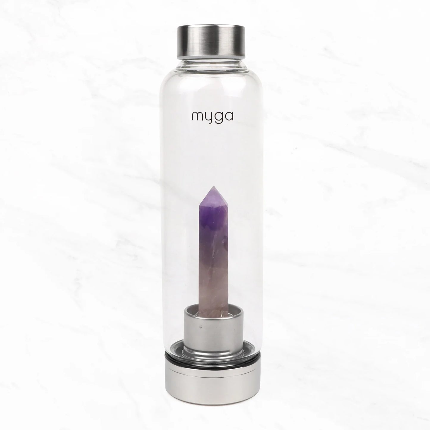 Myga Kristallivesipullo Ametisti; kirkas lasinen vesipullo hopeisin metallikansin ja fasetoitu violetti kristallipiikki sisällä seisoo pystyssä valkoisella taustalla; sylinterinmuotoisessa pullossa sileät pinnat ja kiiltävä viimeistely; sana "myga" painettu yksinkertaisin pienin groteskikirjaimin kylkeen; ei pakkausta, ihmisiä tai käsiä näkyvissä.