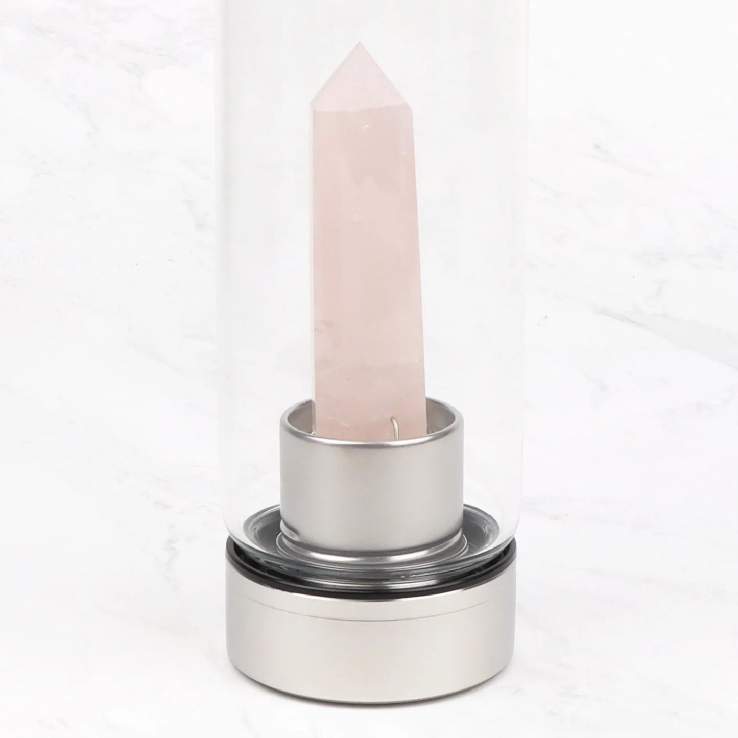 Myga Kristallivesipullo Ruusukvartsi; kirkas lasinen pullo hopeisella jalustalla, sisällä pystyssä seisova terävä ruusukvartsikristalli; pullon pinta on sileä ja kiiltävä; ruusukvartsi on vaaleanpunainen ja terävämuotoinen; ei luettavaa etikettiä tai tekstiä pullossa; tausta on valkoista marmoria, tasaisesti valaistu, kuvattu suoraan edestä ilman ihmisiä.