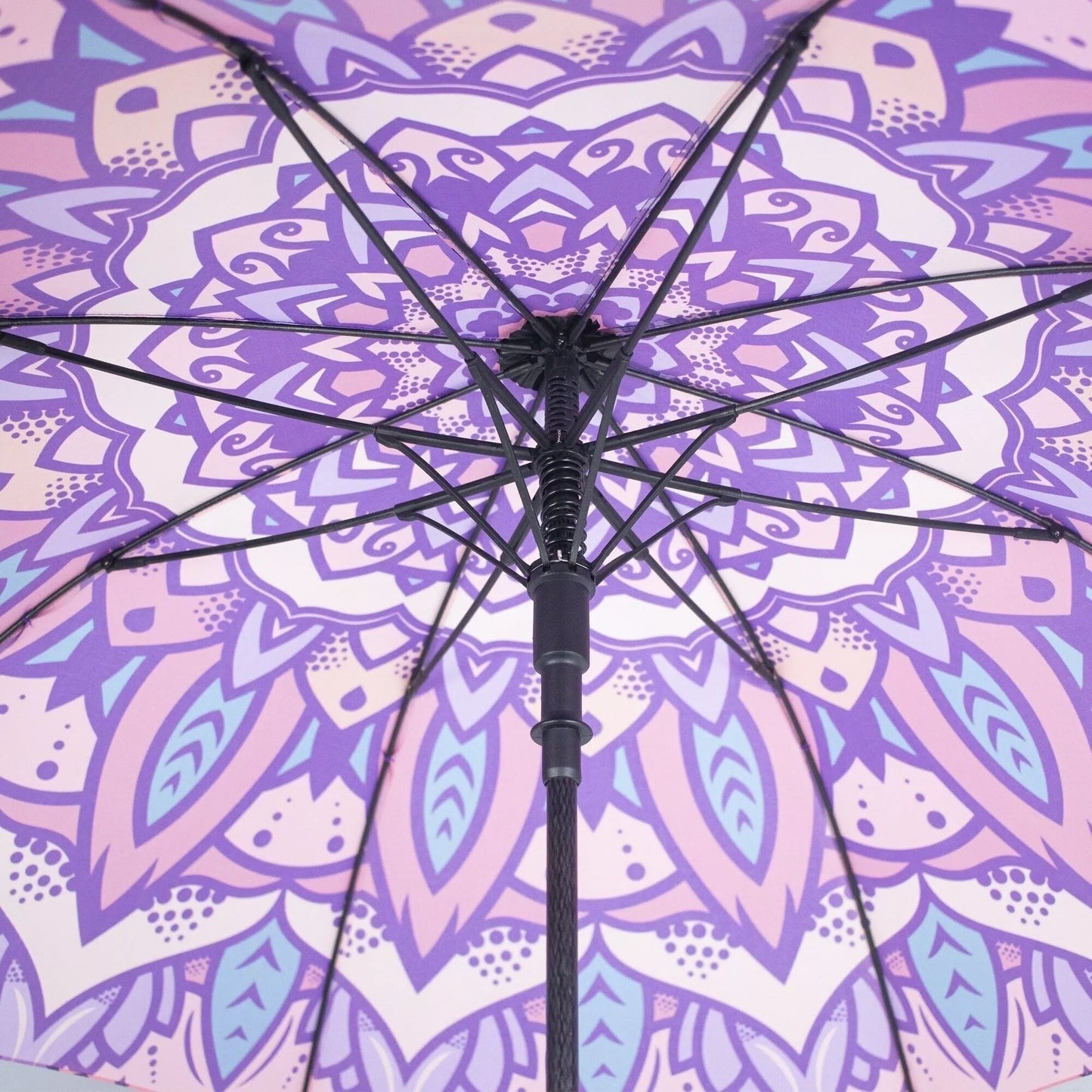 Myga Mandala Sateenvarjo; lähikuva avatun sateenvarjon alapinnasta, violetti ja laventelinvärinen mandala-kuvio geometrisin ja kukkamaisin muodoin; mustat sateenvarjon rungot kehystävät monimutkaista kuviota; katon materiaali mattapinnalla; ei näkyvää pakkausta, etikettitekstiä tai ihmisiä; tausta jää kuvan ulkopuolelle, huomio keskittyy rakenteeseen ja kankaan yksityiskohtiin.