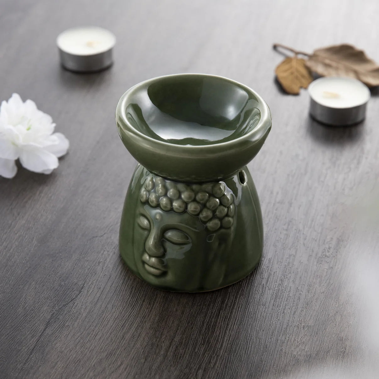 Myga Aromaterapia Öljypoltin; vihreä keraaminen Buddha-päänmuotoinen öljypoltin puupintaisella alustalla, ympärillä lämpökynttilöitä, valkoinen kukka, kuivattuja lehtiä ja rauhoittavia aromaterapia-aiheisia yksityiskohtia; öljypoltimessa mattapinta ja veistetyt kasvonpiirteet; ei pakkausta tai etikettejä näkyvissä; tausta neutraali, esineet tiiviisti aseteltuina; ei ihmisiä tai käsiä kuvassa.