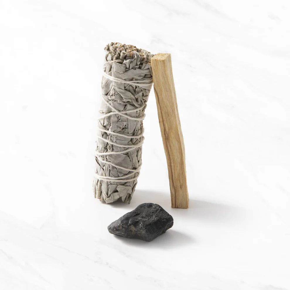 Myga Puhdistusrituaalisarja; nippu kuivattua salviaa, palo santo -tikku ja pieni obsidiaanikivi aseteltuna valkoiselle marmoripinnalle; salvianippu lieriömäinen vaaleanvihrein lehdin valkoisen narun ympäröimänä; palo santo -tikku vaaleanruskea ja suorakaiteen muotoinen näkyvin puun syin; obsidiaani sileä, musta, soikea ja kiiltäväpintainen; ei pakkausta tai etikettejä näkyvissä.