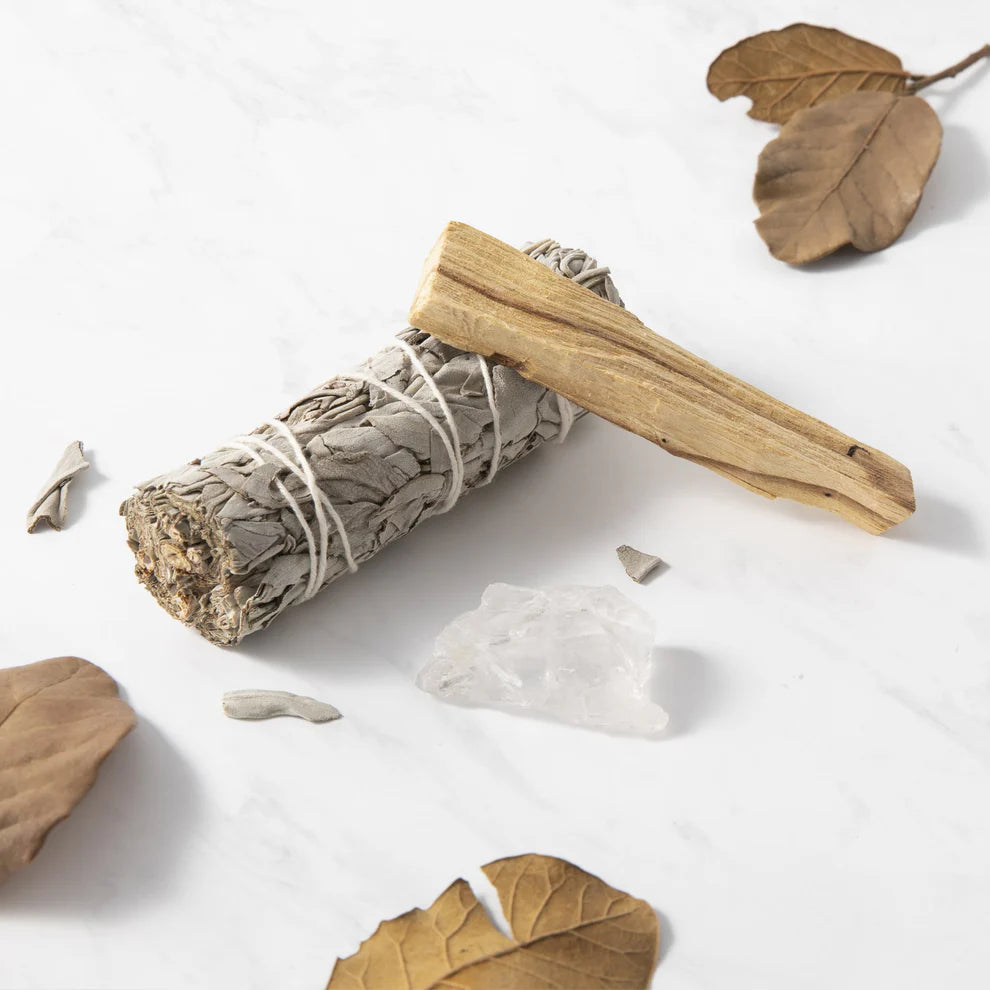 Myga Puhdistusrituaalisarja; nippu kuivattua salviaa, palo santo -tikku, pieni kirkas kvartsikristalli ja kuivia ruskeita lehtiä on aseteltu valkoiselle marmoripinnalle; kaikki esineet avoimina ja luonnonmukaisin tekstuurein, salvia sidottu narulla, palo santo suorakaiteen muotoinen ja vaaleanruskea, lehdet irtonaisia ja mattapintaisia, kristalli kirkas ja monikulmainen; ei etikettejä tai tekstiä; ei ihmisiä tai käsiä; kuvattu suoraan ylhäältä.