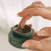 NOBE Forest Elixir® Microbiome Restoring Day & Night Cream: manikyroitu käsi kastaa sormen pyöreään, läpinäkymättömään vihreään purkkiin tekstillä "Forest Elixir Microbiome Restoring Day & Night Cream" valkoisella groteskitekstillä; mattapintainen kansi on irrotettu, paljastaen paksun ruskean voiteen sisällä; kuva on lähikuva, tausta on suurelta osin epätarkka ja neutraalinvärinen.