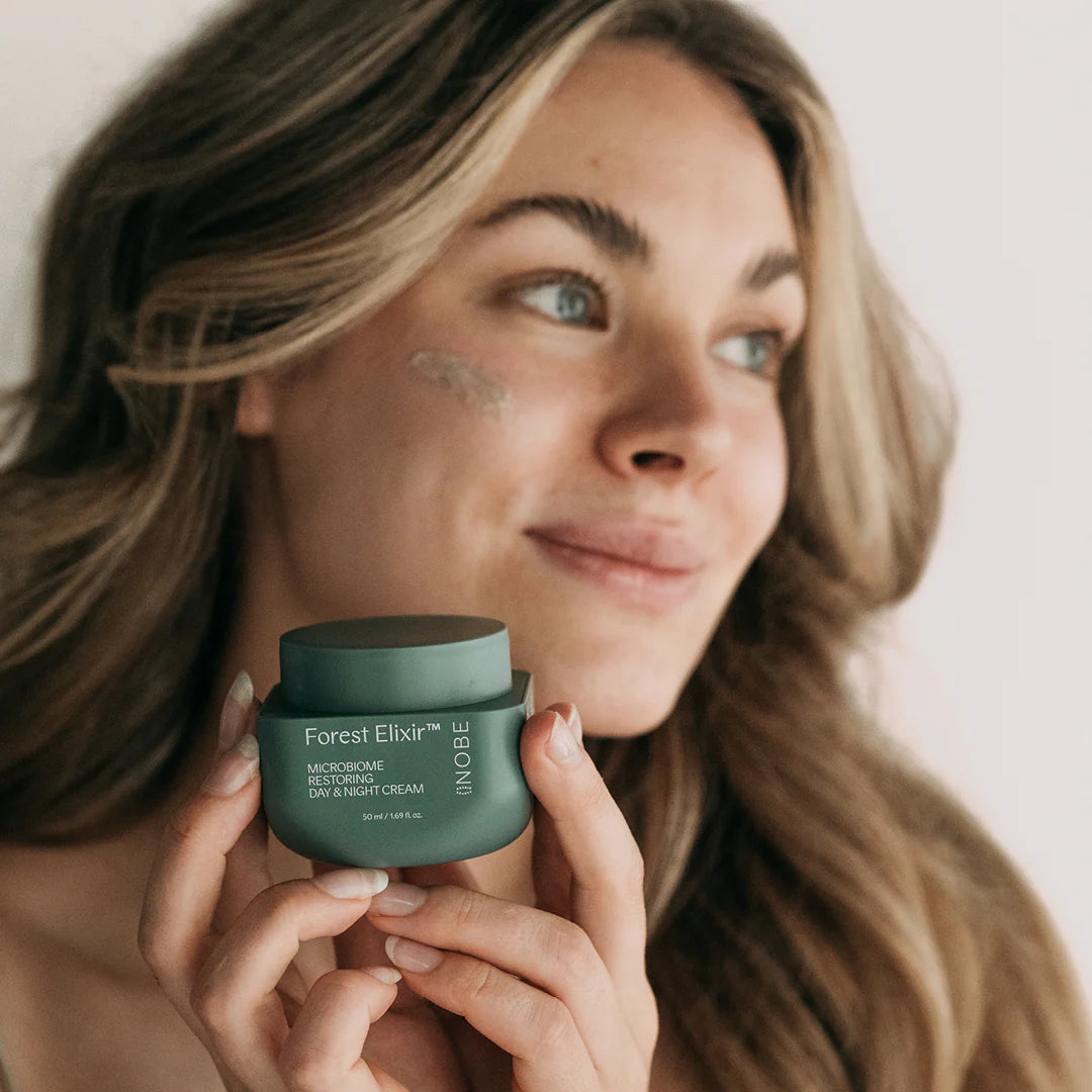 NOBE Forest Elixir® Microbiome Restoring Day & Night Cream: hymyilevä nainen pitelee vihreää purkkia tekstillä "Forest Elixir Microbiome Day & Night Cream" valkoisella kirjoituksella; purkki on sylinterimäinen mattapinnalla ja yhteensopivalla kannella; naisen poskella näkyy voidetta ja hänellä on pitkät aaltohiukset; kuva on lähikuva muotokuvana neutraalia taustaa vasten; kaava tukee ihon mikrobiomia.