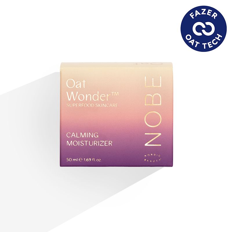 A box of NOBE Oat Wonder Calming Moisturizer, 50 ml, featuring a soothing purple and beige gradient design. The surface is matte with rectangular packaging. The blue Fazer Oat Tech logo is in the top right corner. Typography is clean and sans-serif. No people or hands are present. Photo has a plain background and uses direct lighting.