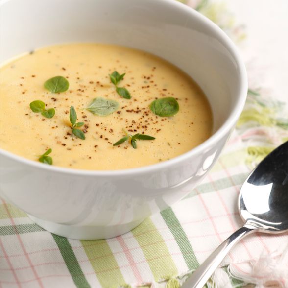 A white bowl of turmeric-infused creamy yellow soup, garnished with green herbs and black pepper, sits on a plaid cloth next to a metal spoon. The bowl is ceramic with a matte finish and round shape. The plaid cloth features red and blue tones as the surface setting. No packaging, labels, text, or people are present in the image. Overhead shot captures all elements clearly.