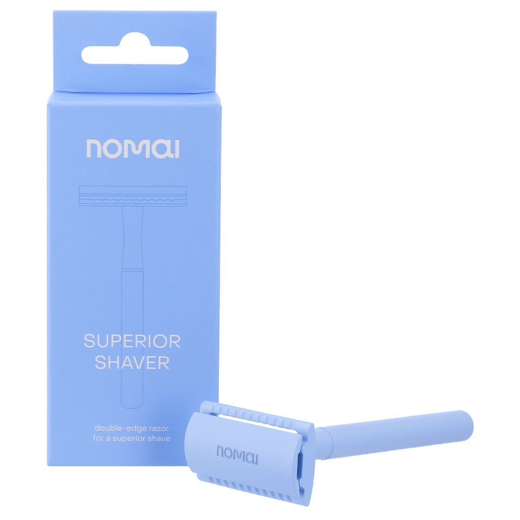 Nomai Superior Shaver; sininen Nomai-partaterä aseteltuna sinisen laatikkonsa eteen, jossa brändinimi ja teksti Superior Shaver kirkkaan valkoisin kirjaimin; terä metallisella pinnalla, suora varsi ja litteä pää; laatikko suorakaiteen muotoinen, mattasininen, valkoinen groteskiteksti; vaalea tausta, ei ihmisiä tai käsiä; lähikuva tuotteesta.