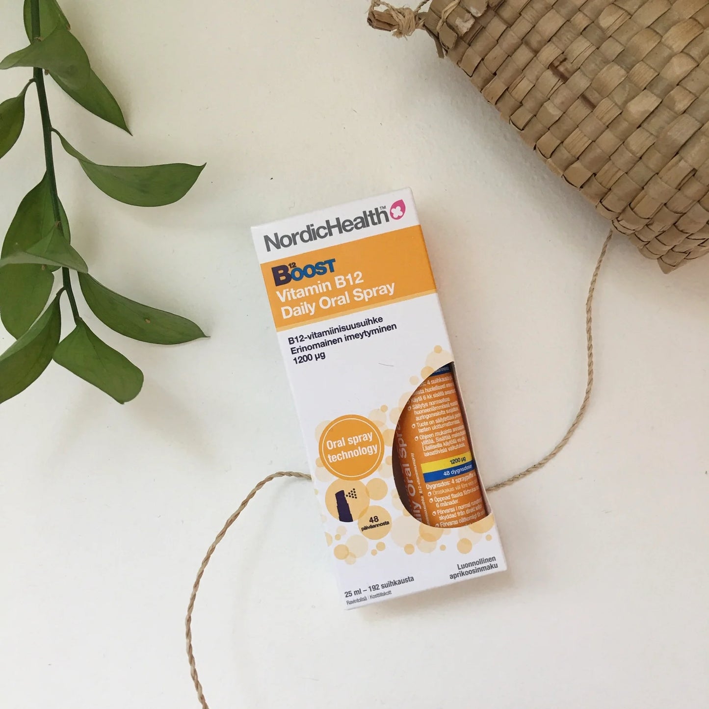 Nordic Health Boost B12-vitamiinisuihke -pakkaus, valkoinen-punainen mattapintainen rasia, lihavoitu groteskiteksti "Vitamin B12 Daily Oral Spray"; vieressä punottu kori ja vihreä lehväoksa