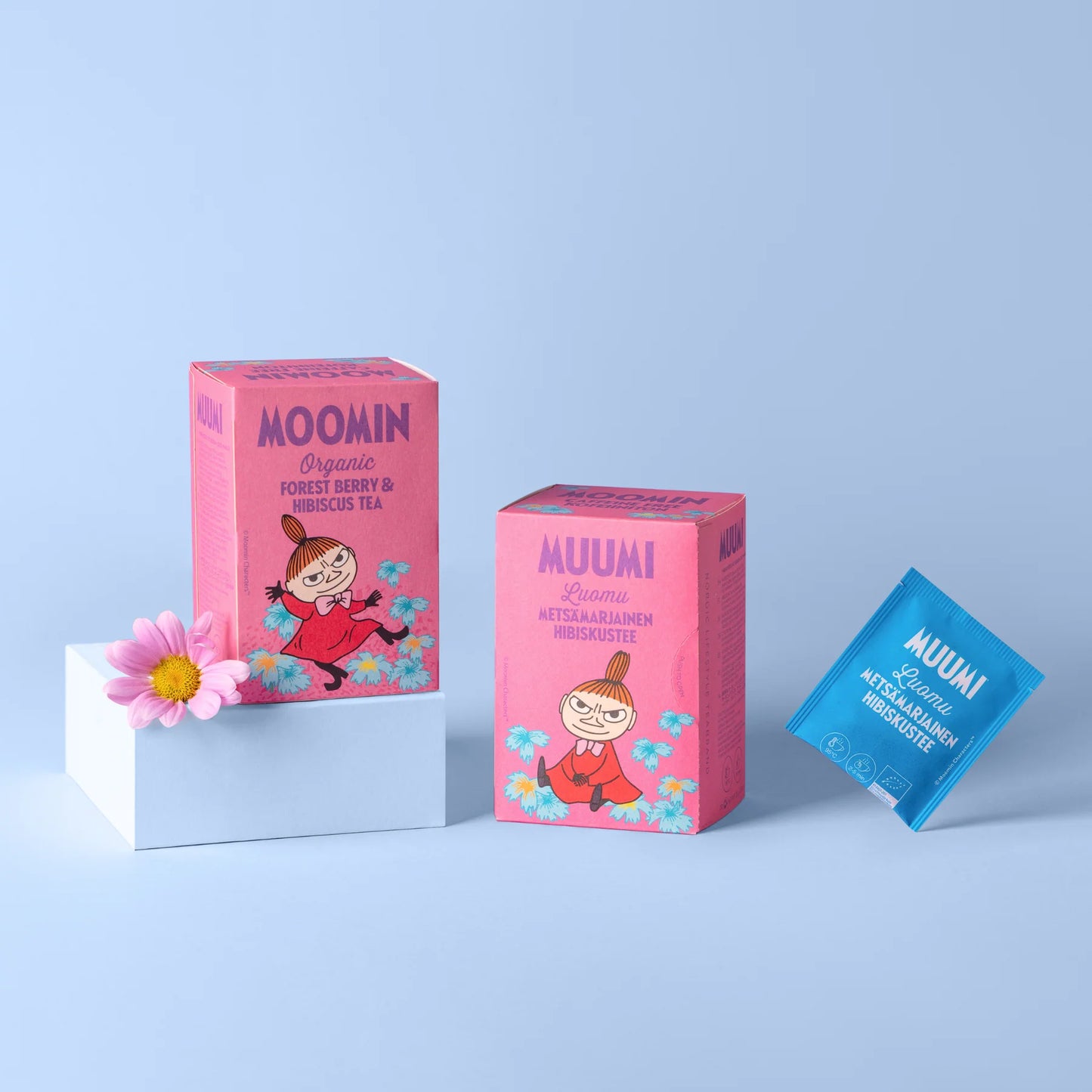 Muumi Luomu Metsämarjainen Hibiskustee: kaksi pinkkiä Muumi-teerasiaa mattapinnalla sarjakuvahahmokuvituksin, aseteltuna sinisen teepussin viereen lihavoidulla valkoisella tekstillä; pinkki kukka lepää tuotteiden vieressä valkoisella suorakulmaisella alustalla; tausta on yhtenäisen vaaleansininen; ei ihmisiä tai käsiä näkyvissä tässä studiotyylisessä tuotekuvassa.