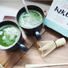 Nordqvist Riku Matcha Teejauhe; kaksi mustaa lieriömäistä mukia täynnä vaahtoavaa vihreää matcha-teetä puisella tarjottimella, vieressä bambuvispilä, vihreä sekoitustikku ja osittain näkyvä matcha-teepakkaus; mukit mattapintaisia kaartuvin kahvoin; hopeinen pakkaus vihrein aksentein; ei ihmisiä tai käsiä.