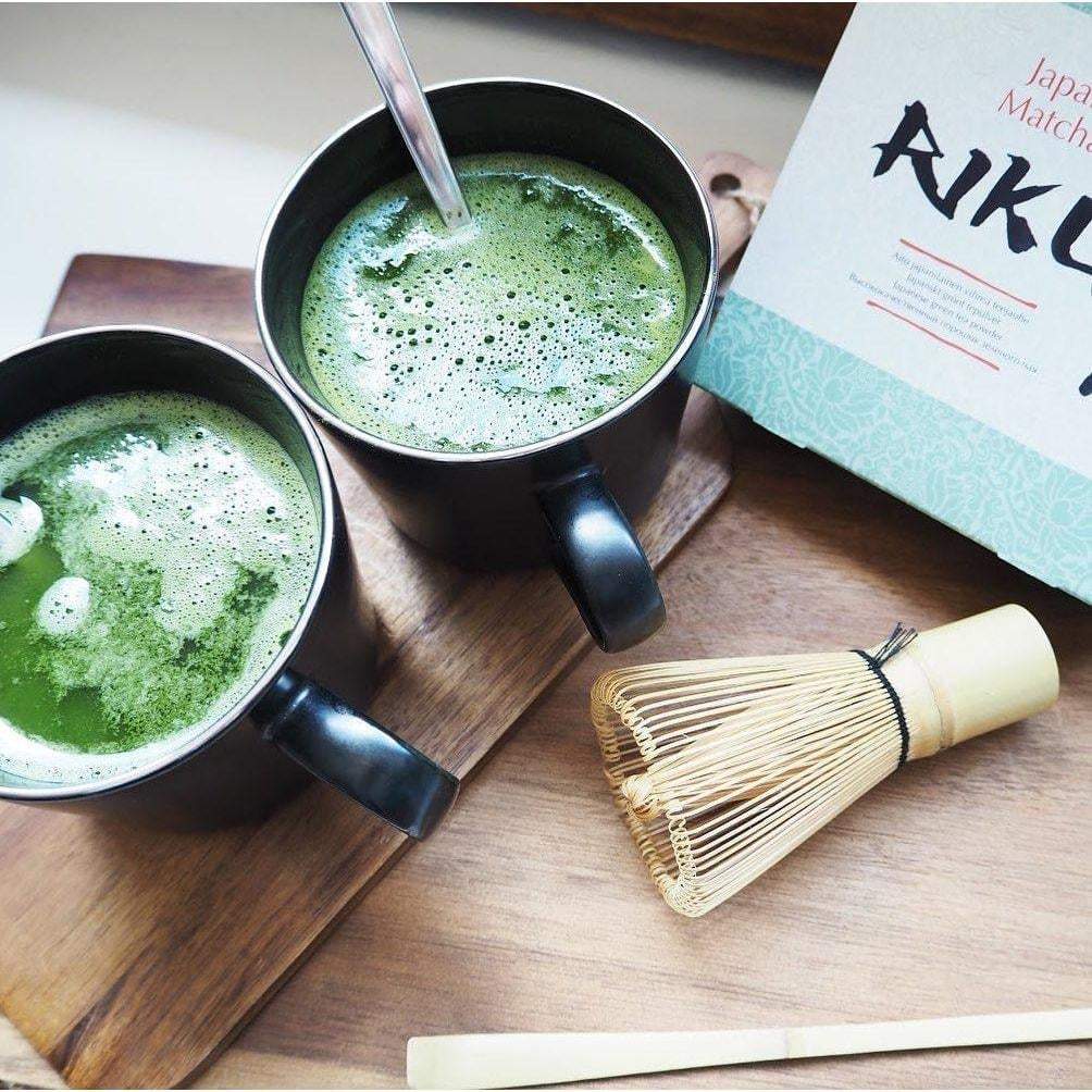 Nordqvist Riku Matcha Teejauhe; kaksi mustaa lieriömäistä mukia täynnä vaahtoavaa vihreää matcha-teetä puisella tarjottimella, vieressä bambuvispilä, vihreä sekoitustikku ja osittain näkyvä matcha-teepakkaus; mukit mattapintaisia kaartuvin kahvoin; hopeinen pakkaus vihrein aksentein; ei ihmisiä tai käsiä.