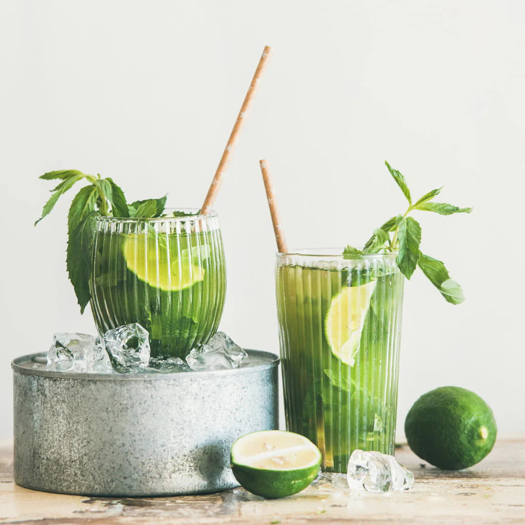 Nordqvist Riku Matcha Teejauhe; kaksi läpinäkyvää lasillista mojitoa jääpaloin, limeviipalein ja mintunlehdin, kummassakin suora muovipilli; kokonainen ja puolikas lime pöydällä lasien vieressä; kirkas, valoisa asetelma; ei ihmisiä tai käsiä kuvassa.